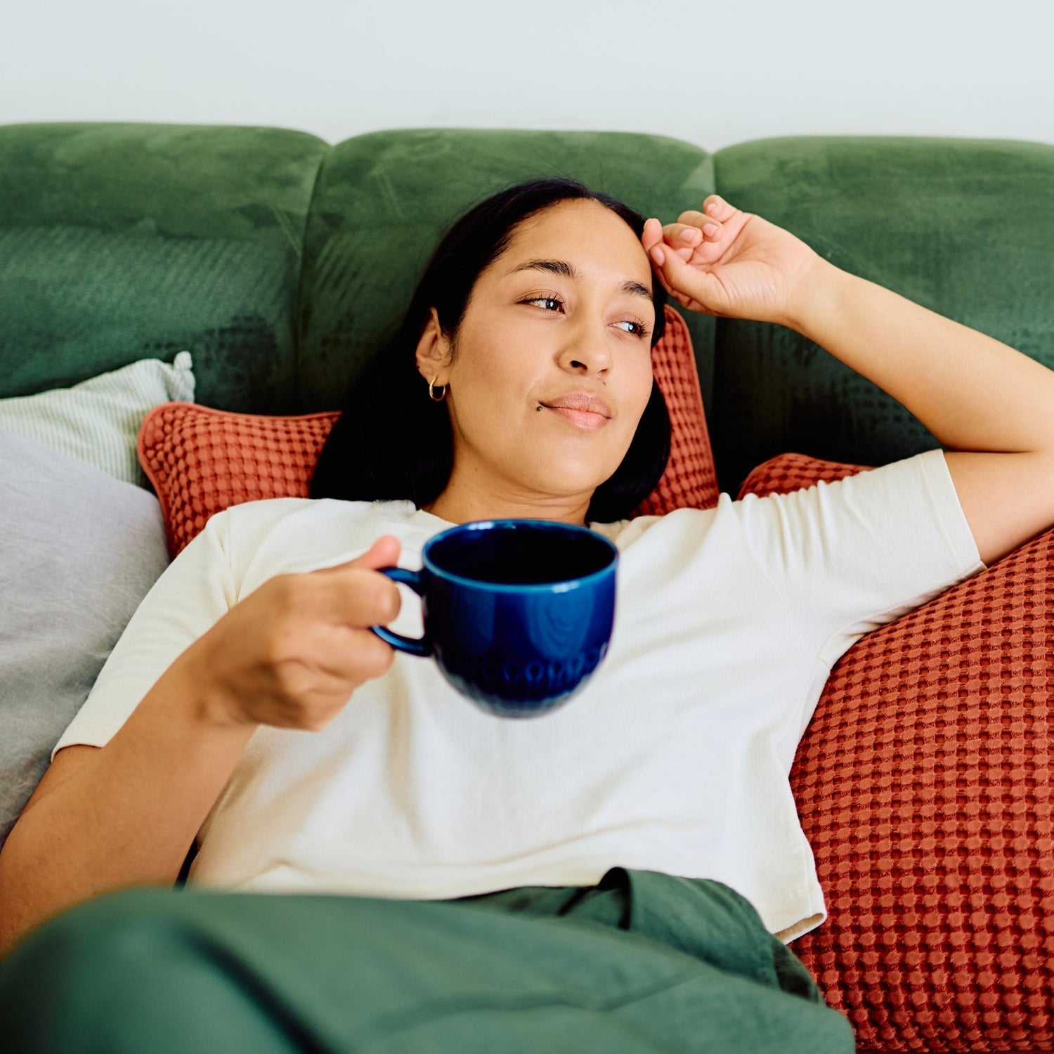 Women laying on a green couch with an orange pillow holding a blug coffee cup - she is resting 