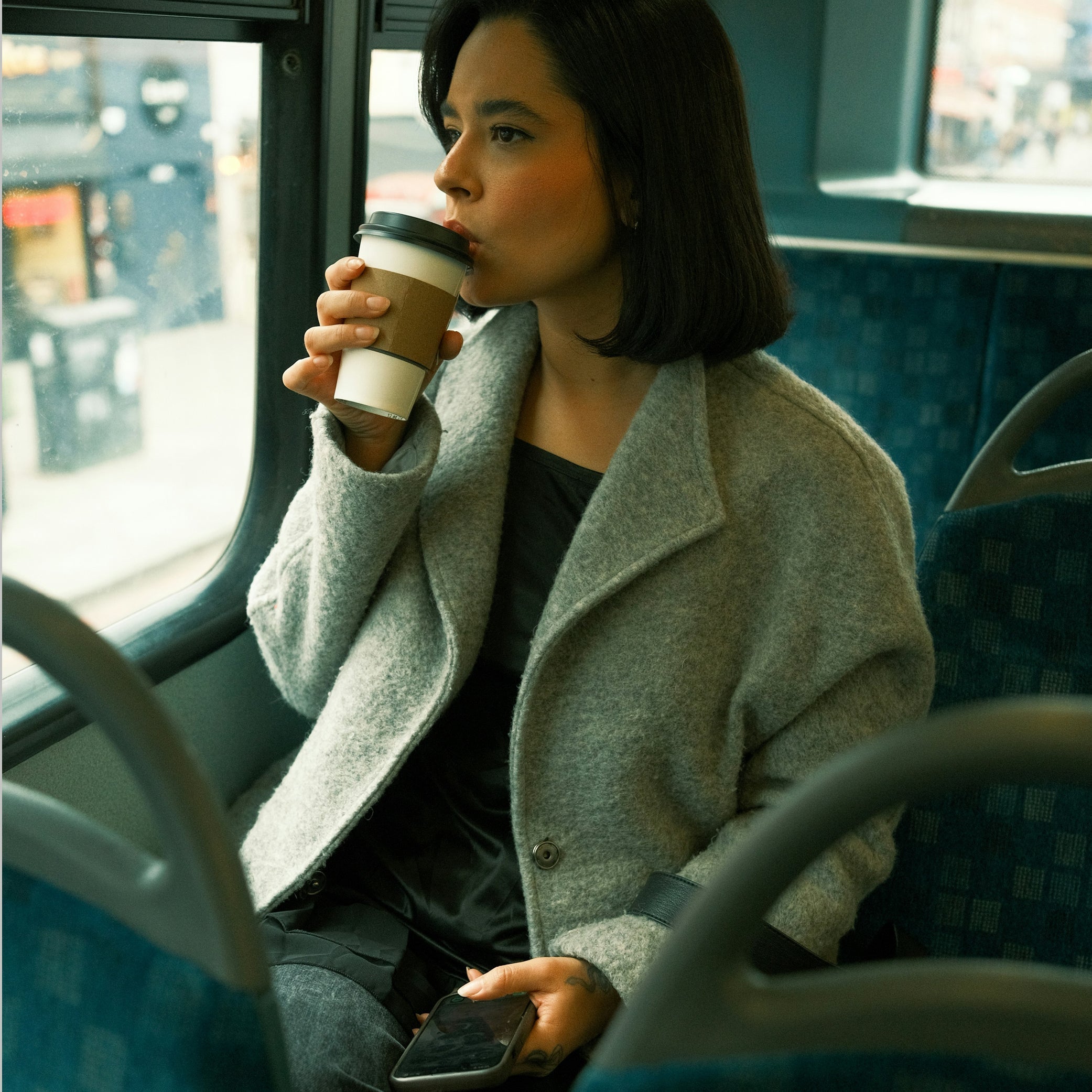 A woman with dark shoulder-length hair sits beside the window on a bus, wearing a grey coat and drinking from a takeaway coffee cup. She looks thoughtfully out at the blurred city street.