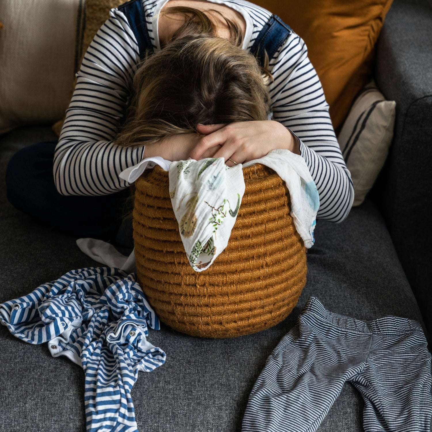 An exhausted mum leans forward with her face hidden in her folded arms, resting on a woven basket of baby laundry on a grey couch